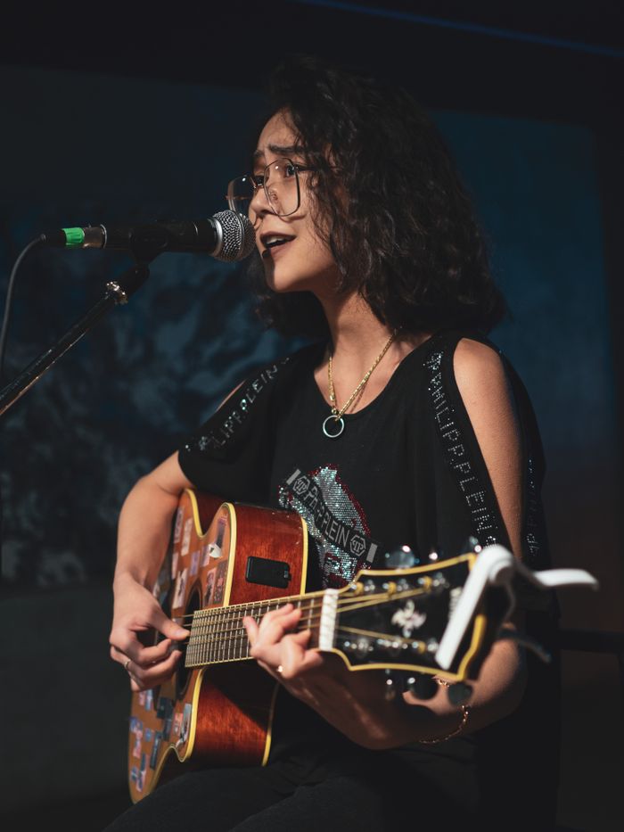 about-01 Young woman with eyeglasses singing and playing acoustic guitar on stage.