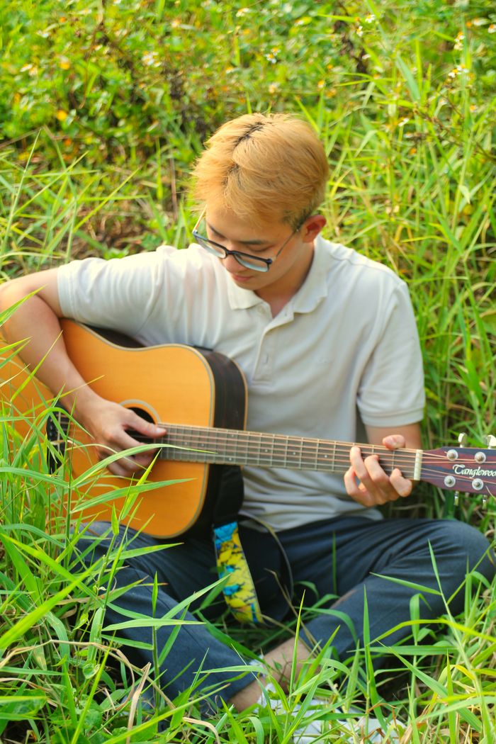 gallery-4 A young man sits outdoors, playing an acoustic guitar in a lush, grassy setting.
